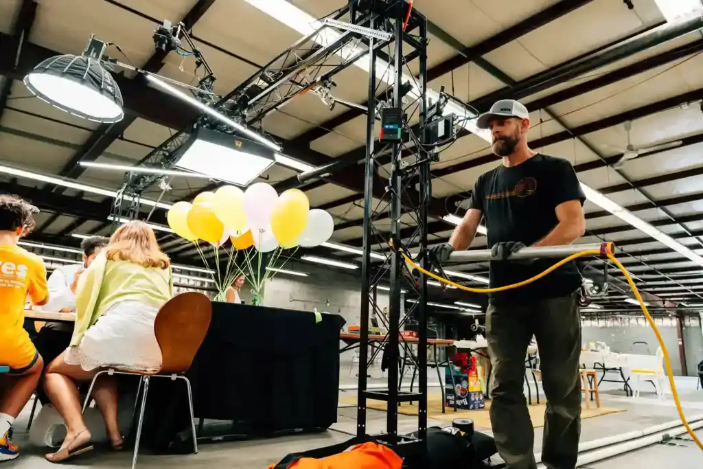 Bearded man in a gray cap and black shirt stands beside a tall metal rig with cables in a workshop, balloons and people at a table in the background.