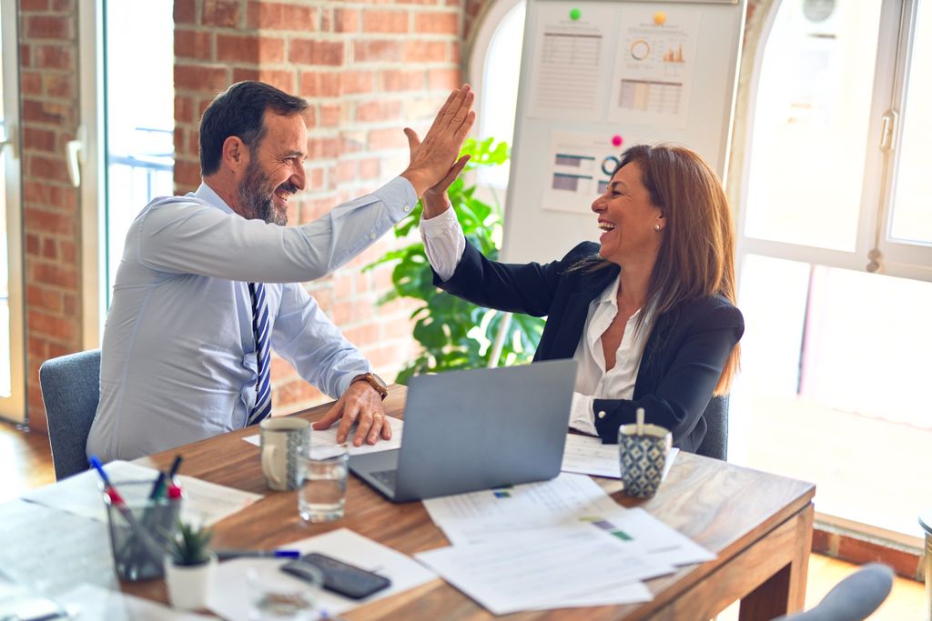 Two colleagues high-fiving over a laptop and printed revenue charts in an office