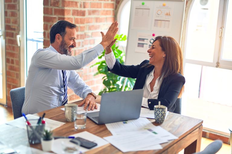 Two colleagues high-fiving over a laptop and printed revenue charts in an office